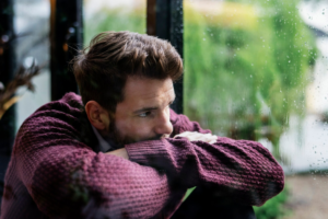 Man looking out of a window, reflecting on his emotional state, representing the isolation men often feel in their mental health struggles